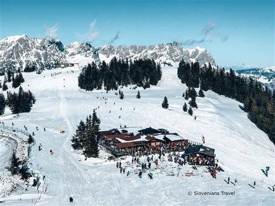 A snowy mountain landscape with skiers and a cozy building. The sunny atmosphere and the surrounding mountains create an inviting winter scene.
