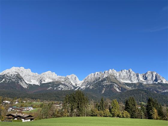 An impressive mountain landscape with snow-capped peaks and a clear blue sky. In the foreground, green meadows and a few houses can be seen.