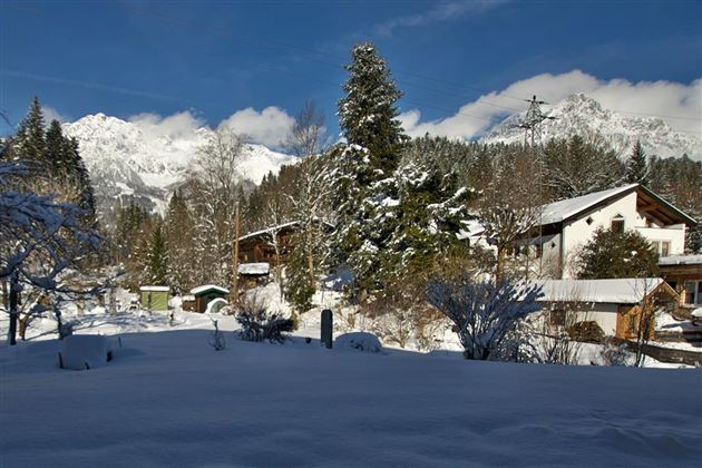 Eine verschneite Landschaft mit traditionellen Chalets und hohen Bergen im Hintergrund. Der Himmel ist klar und blau, was die idyllische Winteratmosphäre unterstreicht.