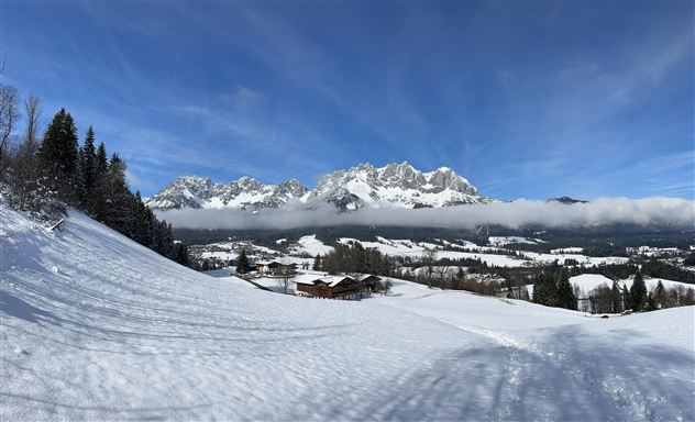 A winter landscape with snow-covered hills and impressive mountains in the background. The sky is clear and blue, creating a calm atmosphere.