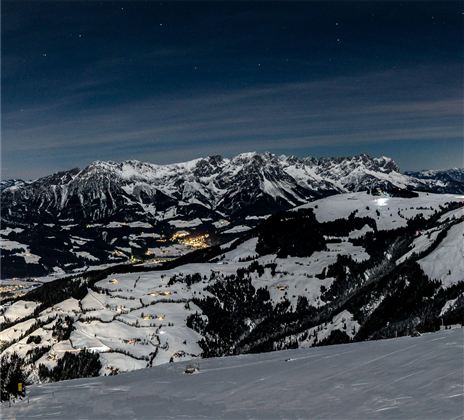 Eine schneebedeckte Berglandschaft unter dem nächtlichen Himmel. In der Ferne leuchten die Lichter eines kleinen Dorfes.