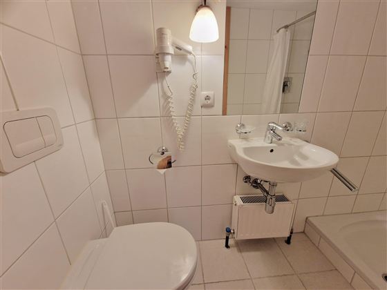 A simple bathroom with white tiles. It features a toilet, a sink, and a shower.