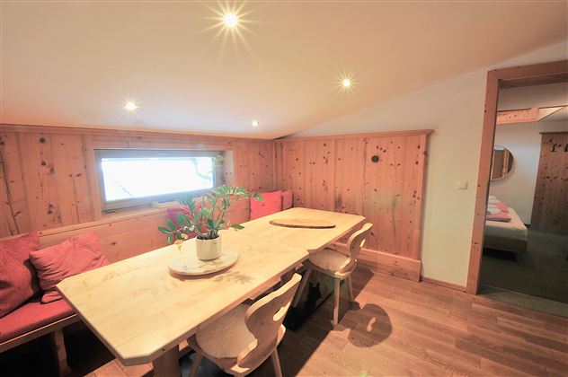 A cozy dining area with a large wooden table and chairs. The walls are made of wood, and there is a windowsill with plants.