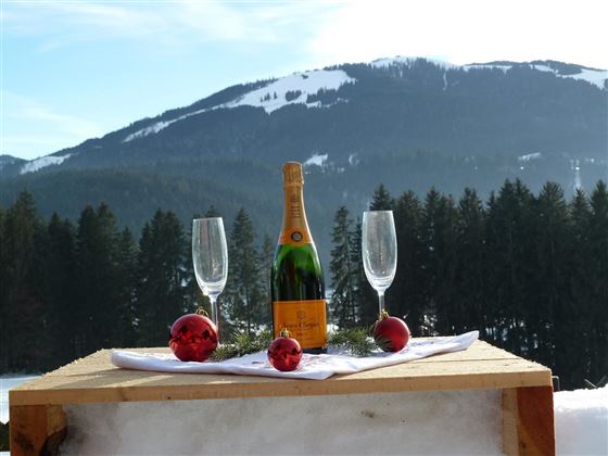 A bottle of sparkling wine is on a wooden table in the snow, surrounded by two glasses and red Christmas ornaments. In the background, snow-covered mountains and forests can be seen.