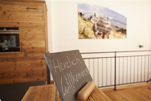 Ein Willkommensschild aus schwarzem Holz steht auf einem Tisch. Im Hintergrund ist ein Bild einer Kuh auf einer Wiese zu sehen.