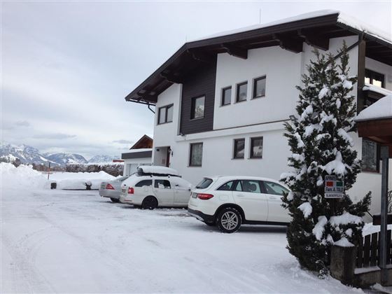 A modern building in the snow with several cars in the driveway. In the background, mountains and a cloudy sky are visible.