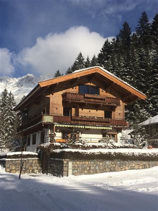 Ein gemütliches Holzhaus im Schnee, umgeben von hohen Tannenbäumen. Der Himmel ist klar und die Berglandschaft ist im Hintergrund sichtbar.