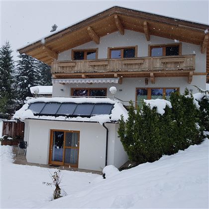A modern house with wooden details, surrounded by snow.  
Flakes gently fall from the sky and the surroundings are peaceful and wintry.