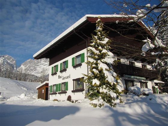 A charming house covered in snow in winter. The surroundings showcase mountains and clear, blue skies.