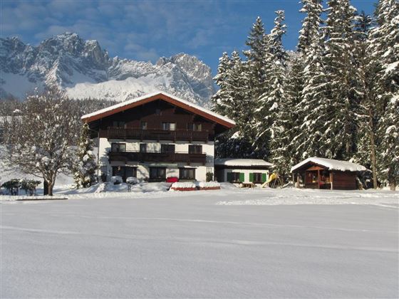 A picturesque house in a snowy landscape, surrounded by tall fir trees and mountains. The sky is clear and blue, creating a serene winter atmosphere.
