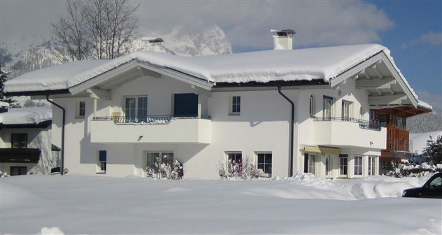 Ein gemütliches Haus im Schnee, umgeben von einer winterlichen Landschaft. Der Himmel ist klar und sonnig, was einen schönen Kontrast zum weißen Schnee bildet.