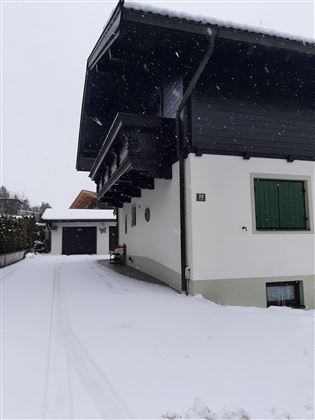 A modern house in a snow-covered landscape. The scene is calm and wintry, with snow-covered paths and gentle snowflakes.