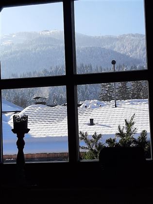 A view from a window of snow-covered roofs and snowy mountains in the background. The sky is clear and blue.