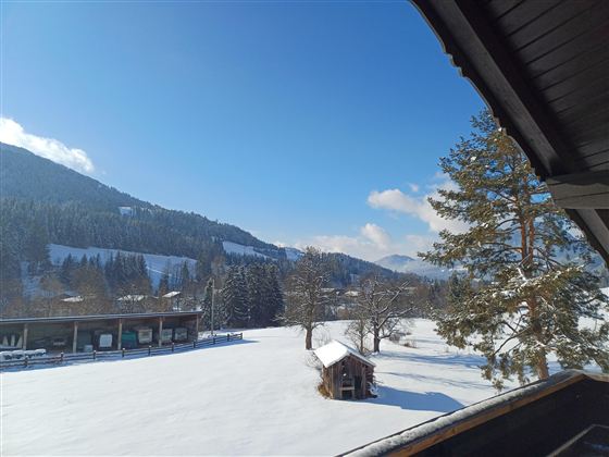 Eine verschneite Landschaft mit Bergen im Hintergrund und einem blauen Himmel. Im Vordergrund steht ein kleiner Holzschuppen und einige Bäume.