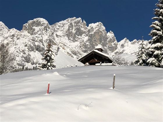 A snowy landscape with a small wooden house and tall mountains in the background. The sky is clear and blue.