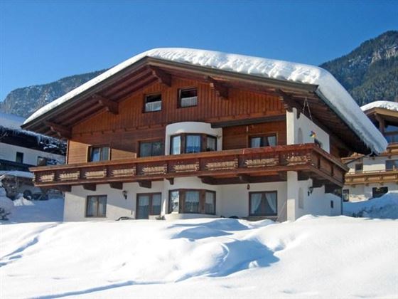 Ein charmantes Holzhaus mit einem schneebedeckten Dach. Die Umgebung ist von weißem Schnee und Bergen umgeben.