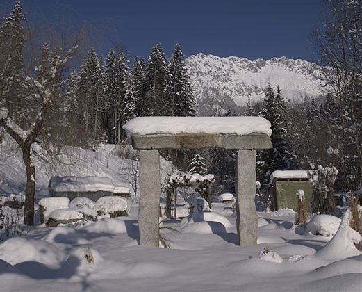 Ein winterlicher Garten mit schneebedeckten Steinen und einer Landschaft aus Bäumen im Hintergrund. Die Berge sind klar und blau am Himmel zu sehen.