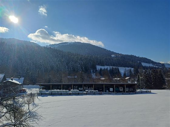 Eine verschneite Landschaft mit Bergen im Hintergrund und einem Gebäude am Rande eines zugefrorenen Sees. Der Himmel ist klar und sonnig.