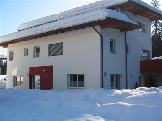 Ein modernes, weißes Haus mit rotem Eingangsbereich, umgeben von schneebedecktem Gelände. Die Dachkante ist mit Schnee bedeckt, und es ist ein klarer Himmel zu sehen.