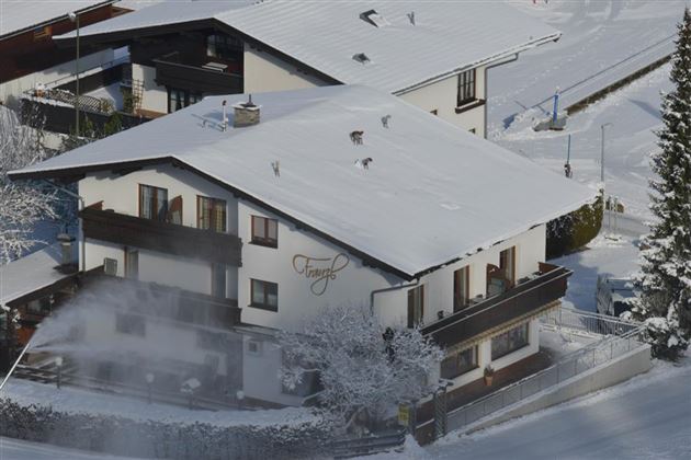Ein gemütliches Haus, bedeckt mit frischem Schnee, in einer winterlichen Landschaft. Im Hintergrund sind Bäume und verschneite Dächer zu sehen.