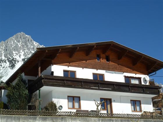 A charming mountain house with a wooden porch and large windows. Majestic mountains are visible in the background.