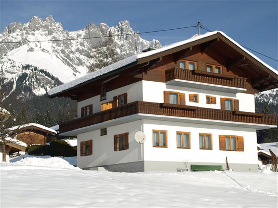 Ein schönes, zweigeschossiges Chalet in den Alpen, umgeben von Schnee. Im Hintergrund erheben sich majestätische Berge unter einem klaren Himmel.