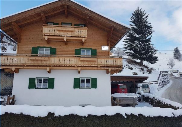 Ein gemütliches Holzhaus im Schnee mit grünen Fensterläden. Umgeben von einer winterlichen Landschaft und Tannenbäumen.