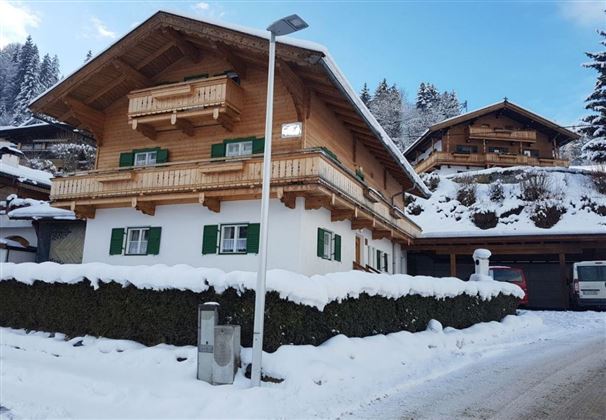 Ein charmantes Chalet im Winter mit weißem Schnee und grünen Fenstern. Im Hintergrund sind weitere Holzhäuser und schneebedeckte Bäume zu sehen.