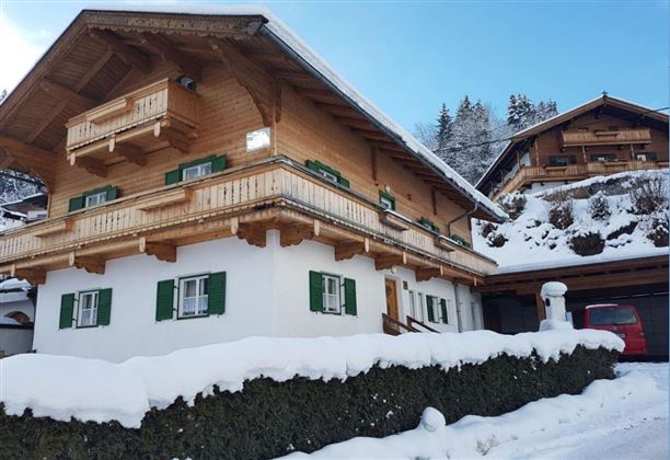 Ein traditionelles chaletartiges Haus im Schnee mit grünen Fensterläden. Die umliegende Landschaft ist winterlich und malerisch.