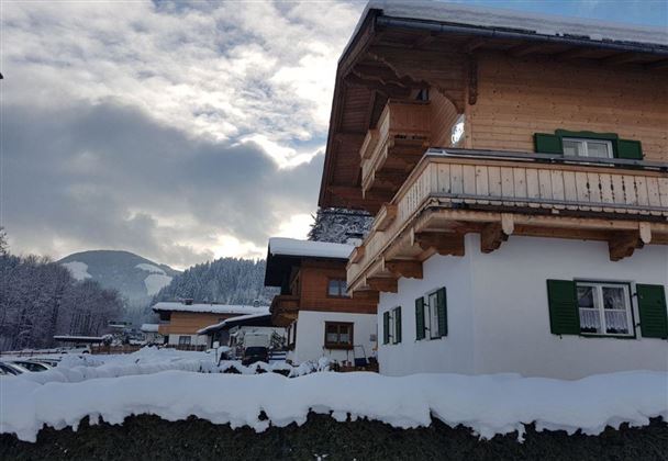 Ein malerisches Winterdorf mit schneebedeckten Dächern und einem hügeligen Hintergrund. Die Gebäude haben traditionelle Holzverzierungen und grüne Fensterläden.