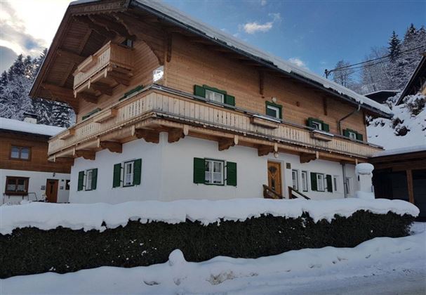Ein charmantes Holzhaus im Winter, umgeben von Schnee. Die grünen Fensterläden und der Balkon verleihen dem Gebäude eine alpine Note.