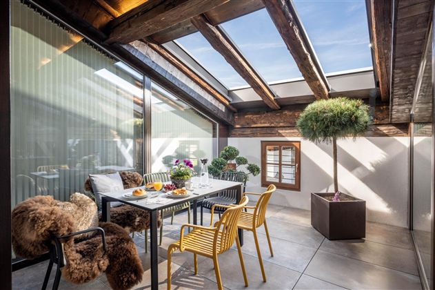 A bright, modern conservatory with a large dining table and comfortable chairs. Green plants and plenty of daylight create an inviting atmosphere.