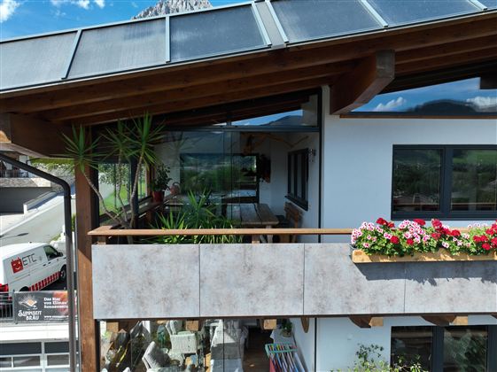Ein schönes Holzhaus mit einer Terrasse, Wintergarten die mit Blumen dekoriert ist. Im Hintergrund sind Berge und ein blauer Himmel zu sehen.