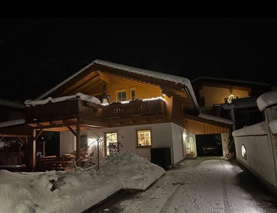 Een gezellig huis in de sneeuw, verlicht door warme lichten. De omgeving is kalm en winters.