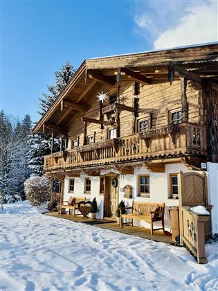 A cozy wooden house in winter, surrounded by snow and fir trees. The balconies are decorated with Christmas ornaments.
