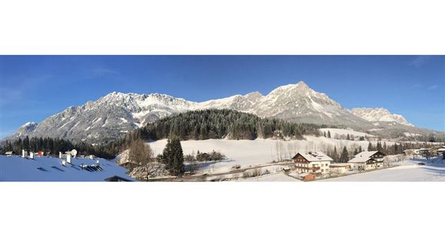 An impressive mountain landscape in winter with snow-covered peaks. In the foreground, wide fields and some buildings can be seen.