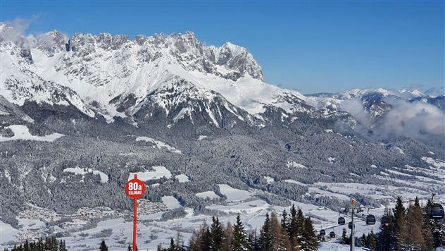 Eine winterliche Berglandschaft mit schneebedeckten Gipfeln und einem klaren blauen Himmel. Im Vordergrund steht ein Hinweiszeichen mit einer Geschwindigkeitsbezeichnung.