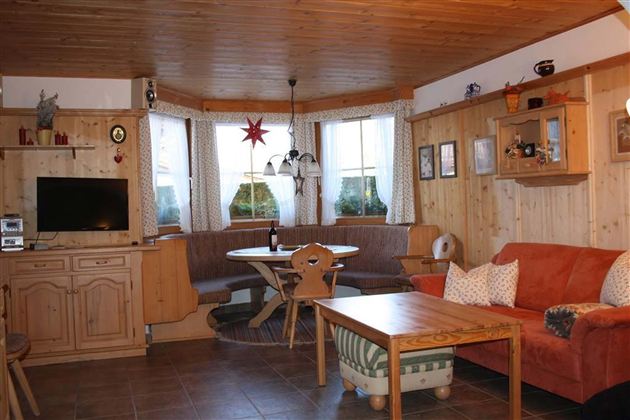 Ein gemütliches Wohnzimmer mit Holzverkleidung, einem roten Sofa und einem runden Esstisch. Große Fenster lassen viel Licht herein und schaffen eine einladende Atmosphäre.