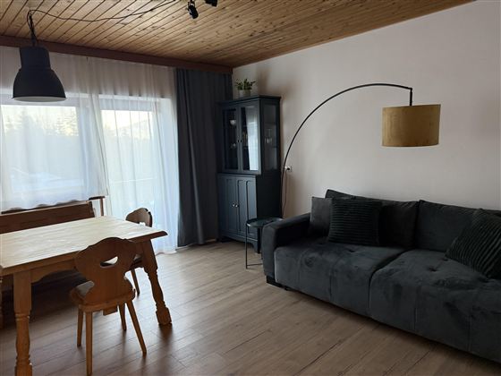 Ein gemütliches Wohnzimmer mit einem grauen Sofa und einem Holz-Esszimmertisch. Große Fenster sorgen für viel Tageslicht und eine entspannte Atmosphäre.