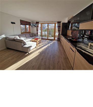 Ein modernes Wohnzimmer mit großen Fenstern und viel Tageslicht. Es gibt ein bequemes Sofa und eine Küchenzeile.