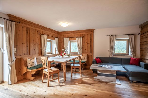 Een gezellige woonkamer met houten wanden en een eettafel. Aan de rechterzijde bevindt zich een moderne bank en grote ramen laten veel licht binnen.