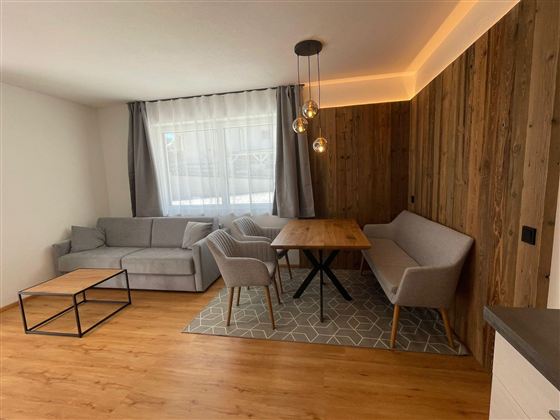 Ein modernes Wohnzimmer mit einem grauen Sofa, einem Holztisch und hellen Wänden. Die große Fensterfront lässt viel Licht herein und sorgt für eine freundliche Atmosphäre.