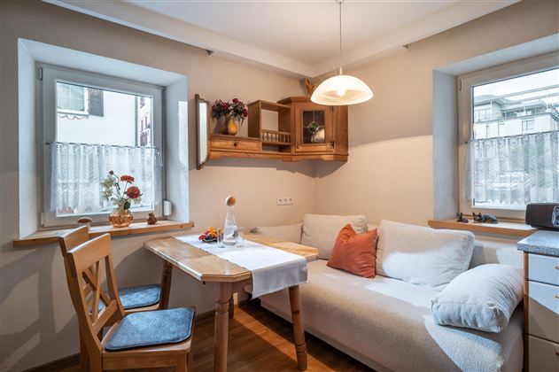 Ein gemütlicher Raum mit einem Tisch, Stühlen und einer Couch. Das Licht fällt durch die Fenster und es gibt dekorative Pflanzen.