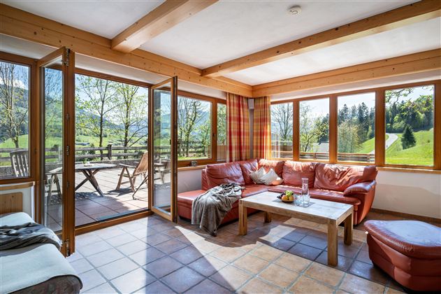 Ein helles Wohnzimmer mit großen Fenstern und einer bequemen roten Couch. Der Blick ins Freie zeigt eine grüne Landschaft und eine Terrasse.