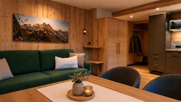 Ein modernes Wohnzimmer mit einer grünen Couch und einem Holztisch. Im Hintergrund sind Küchenschränke und ein schönes Bergbild an der Wand zu sehen.