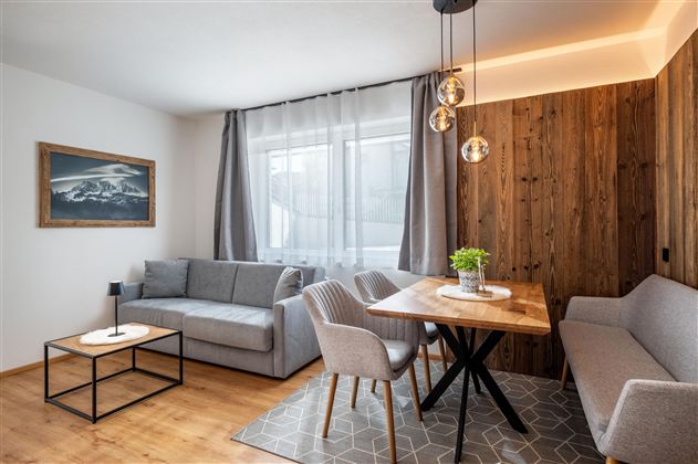 Ein modernes Wohnzimmer mit einem grauen Sofa und einem Holztisch. Große Fenster sorgen für viel Tageslicht und eine angenehme Atmosphäre.