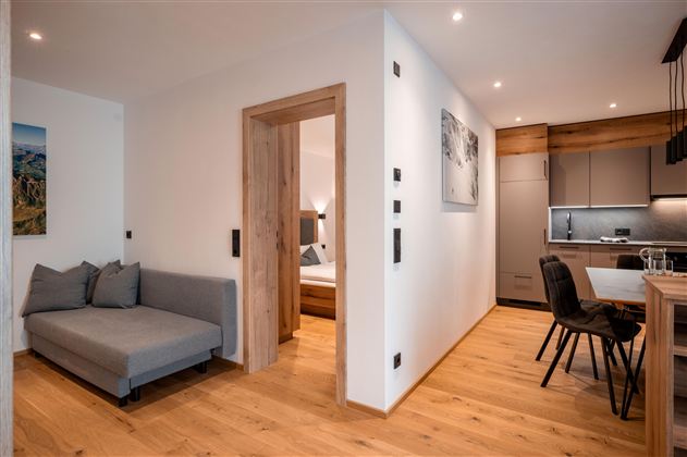 Ein modernes Wohnzimmer mit einem grauen Sofa und Holzboden. Die Umgebung ist hell und freundlich, mit einer Küche im Hintergrund.