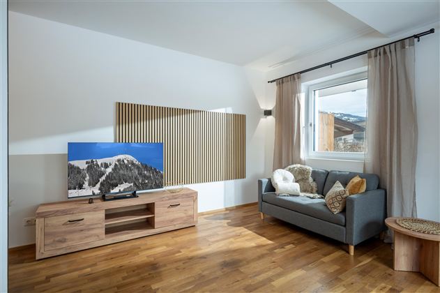 Ein modernes Wohnzimmer mit einem grauen Sofa und einem Holzschrank. Der Fernseher zeigt eine Berglandschaft und große Fenster sorgen für natürliches Licht.