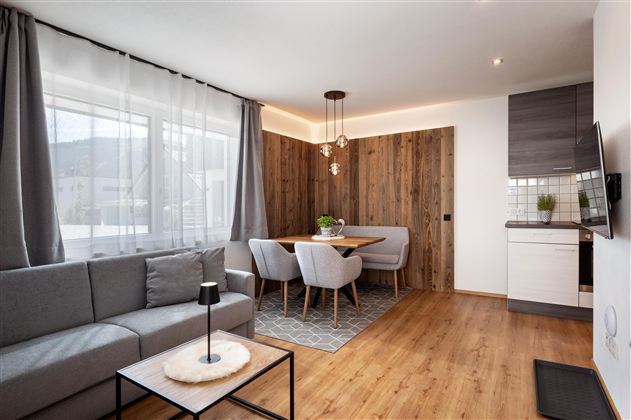 Ein modernes Wohnzimmer mit einem grauen Sofa und einem Esstisch für vier Personen. Große Fenster lassen viel Licht herein und sorgen für eine einladende Atmosphäre.