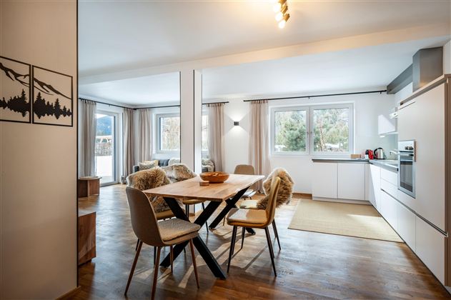 Ein helles Wohnzimmer mit einem großen Esstisch und Stühlen. Der Raum ist gemütlich eingerichtet mit Fenstern und weichen Vorhängen.